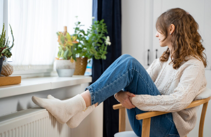 Frau sitzt auf einem Stuhl und stellt ihre Füße auf den Heizkörper, während sie aus dem Fenster blickt.
